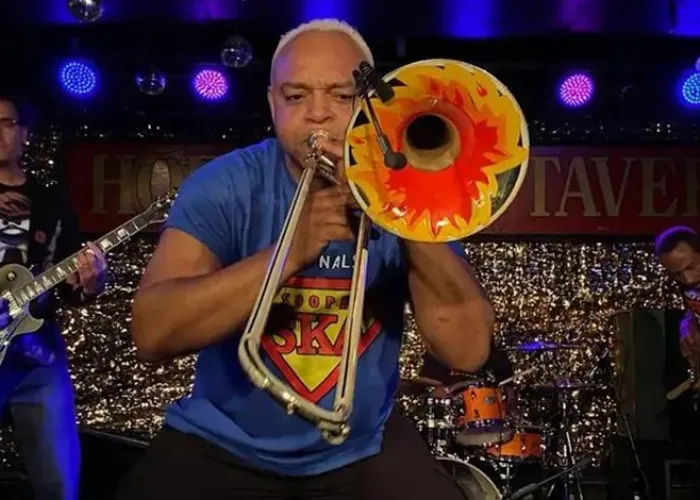 Band member playing colourful trombone in foreground, two band members on guitar and one on drums in background