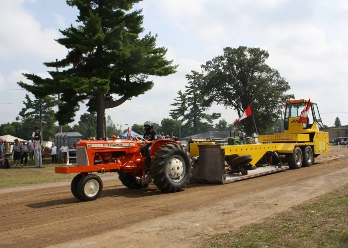 Red tractor pulling yellow machinery decorated with two canadian flags, trees and crowd watching in background